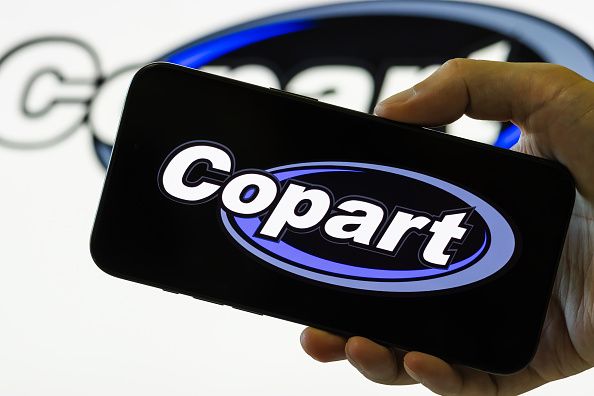 n this photo illustration, a person holds a smartphone displaying the logo of Copart Inc. (NASDAQ: CPRT), an American company specializing in online vehicle auctions and remarketing services, with the company's branding visible in the background on August 28, 2025 in Chongqing, China. (Photo illustration by Cheng Xin/Getty Images)