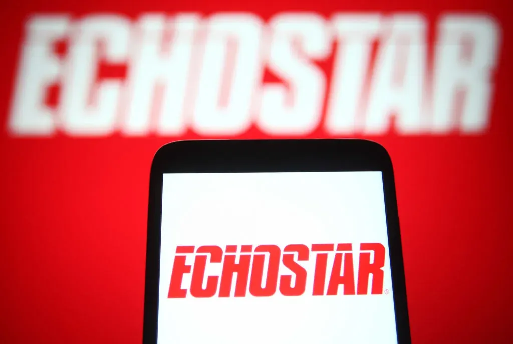 In this photo illustration an EchoStar Corporation logo is seen on a smartphone and a pc screen. 