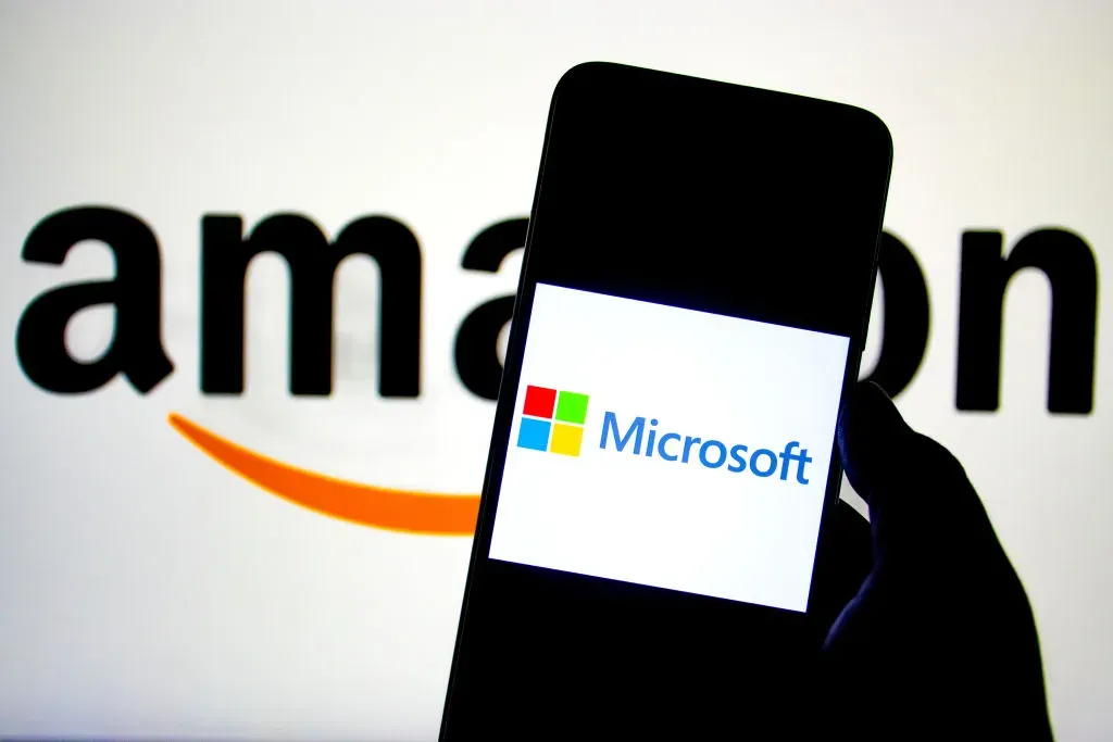 In this photo illustration, a Microsoft logo seen displayed on a smartphone with an Amazon Logo in the background.