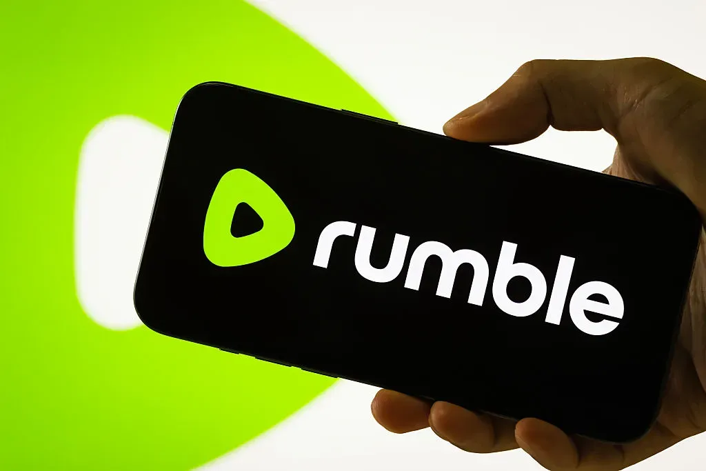  In this photo illustration, a person holds a smartphone displaying the logo of Rumble Inc. 