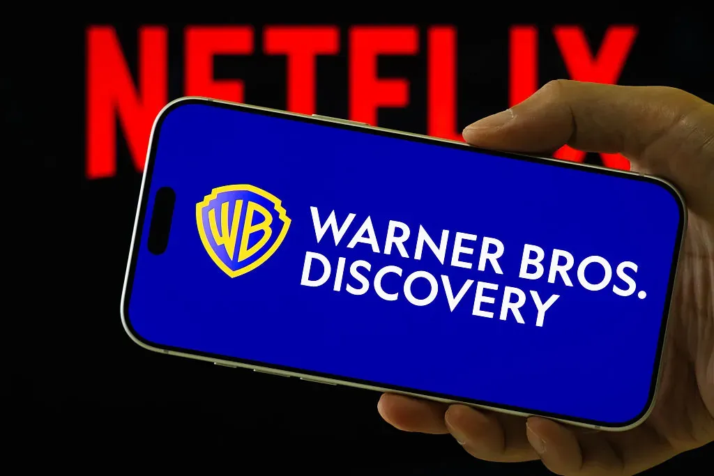 In this photo illustration, a smartphone displays the logo of Warner Bros. Discovery (NASDAQ: WBD) in front of a blurred red Netflix logo.