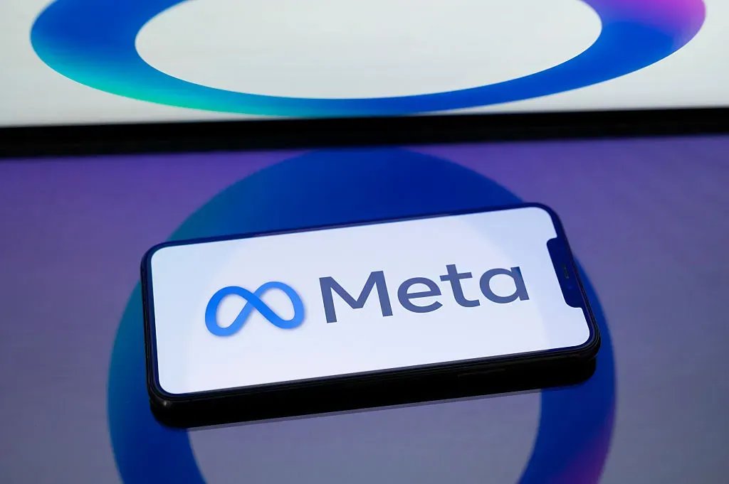 The Meta logo displays on the screen of a smartphone placed on a surface reflecting the circular Meta AI logo.