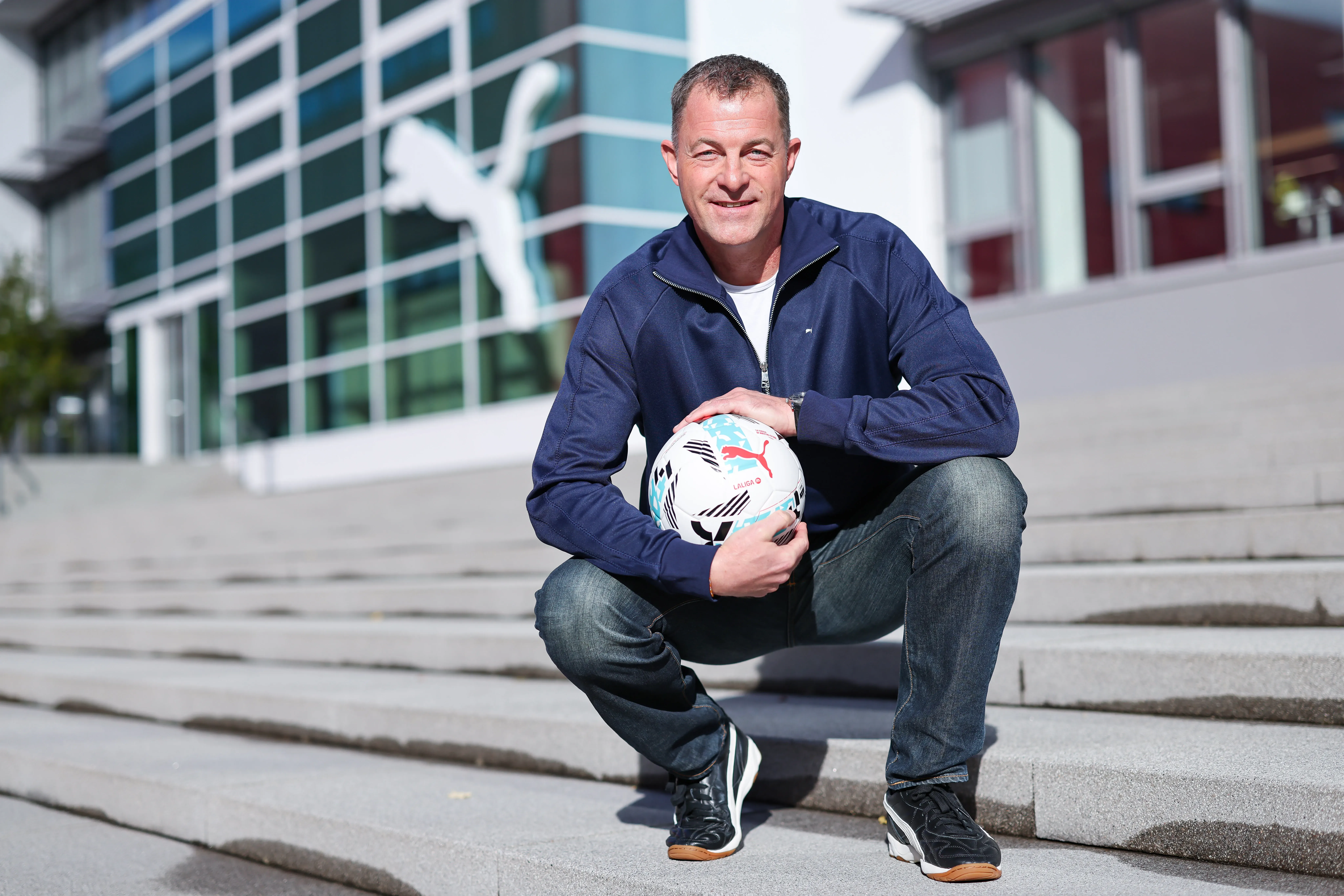Arthur Hoeld, CEO of Puma, kneels with a LaLiga soccer, the Orbita, in front of a large Puma logo at the company headquarters.