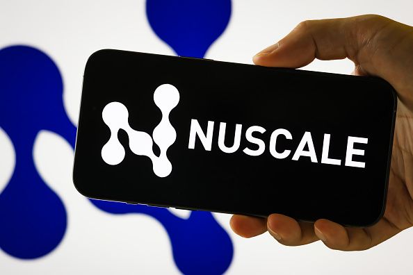 In this photo illustration, a person holds a smartphone displaying the logo of NuScale Power Corporation (NYSE: SMR), a company specializing in small modular nuclear reactor (SMR) technology, in front of a screen showing the company's abstract molecular-style brand icon on August 7, 2025 in Chongqing, China. (Photo illustration by Cheng Xin/Getty Images)