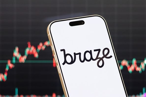 a smartphone displays the logo of Braze, Inc. (NASDAQ: BRZE), an American customer engagement and marketing automation software company, in front of a screen showing the company's latest stock market chart on May 25, 2026 in Shenzhen, Guangdong Province, China. (Photo illustration by Cheng Xin/Getty Images)