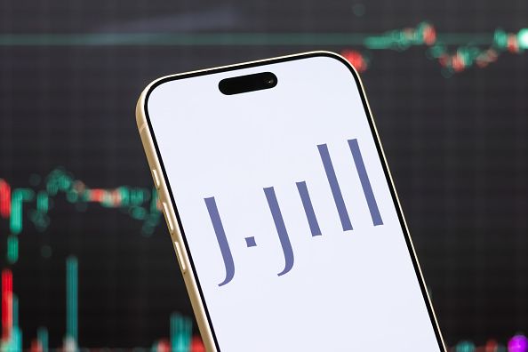 In this photo illustration, a smartphone displays the logo of J.Jill, Inc., an American women's apparel retailer offering clothing, footwear and accessories through retail stores, catalogs and e-commerce channels, in front of a screen showing the company's latest stock market chart on June 13, 2026 in Shenzhen, Guangdong Province, China. (Photo illustration by Cheng Xin/Getty Images)