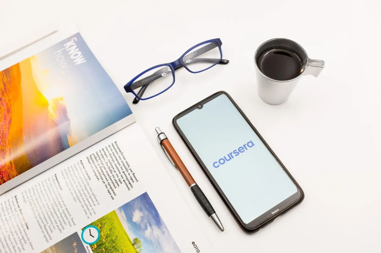 In this photo illustration a Coursera logo seen displayed on a smartphone screen on a desk next to a cafe, a pen, glasses and a magazine in Athens, Greece on June 15, 2022.