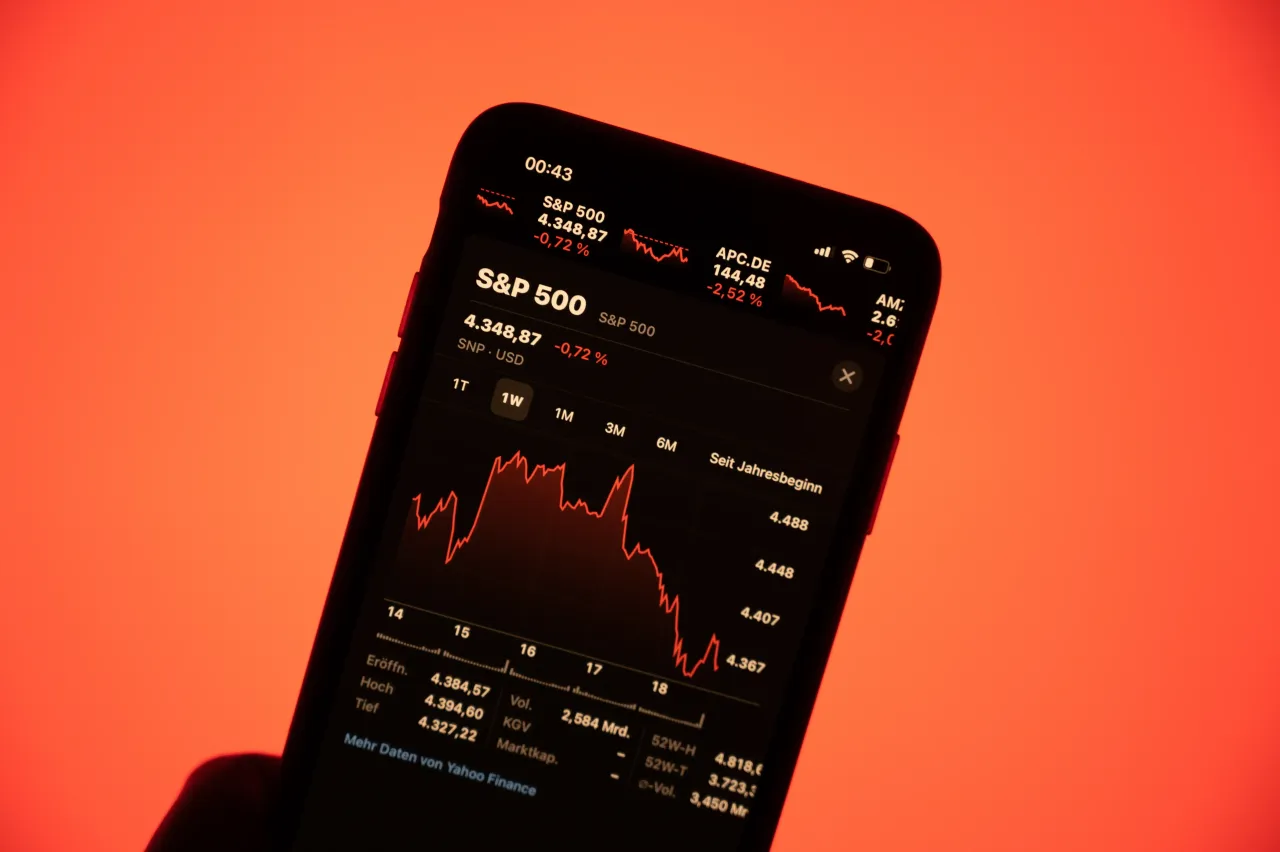 Stock prices of the S&P500 are shown on a smartphone against a red background. 