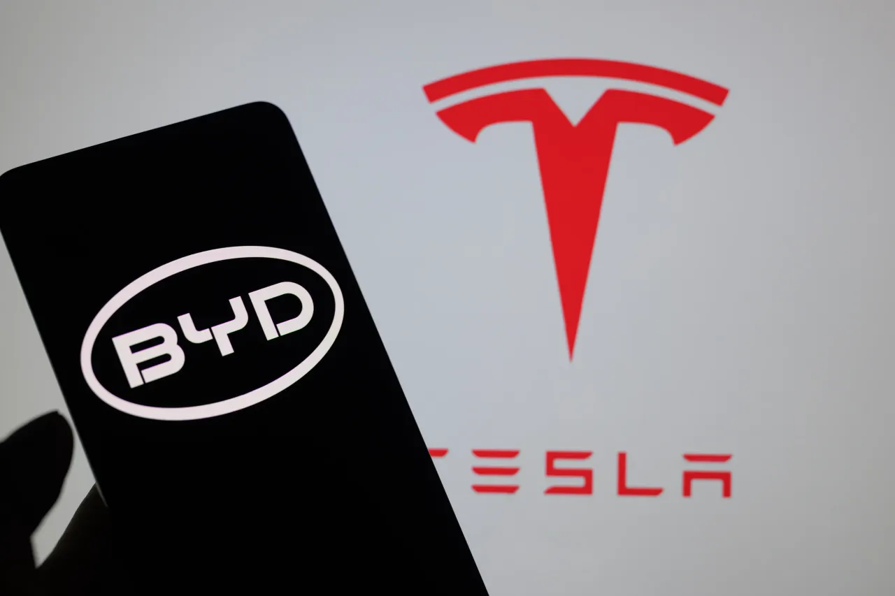 The BYD logo is being displayed on a smartphone with the Tesla logo visible in the background. (Photo by Jonathan Raa/NurPhoto via Getty Images)