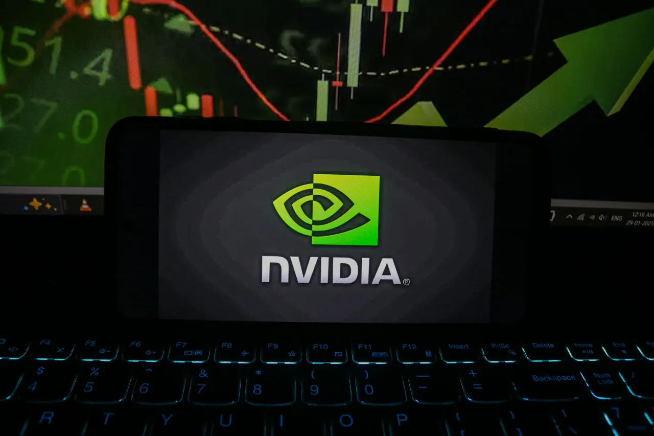 The Nvidia logo appears on a mobile phone, and a stock market graph is on a laptop screen in the background. (Photo by Firdous Nazir/NurPhoto via Getty Images)