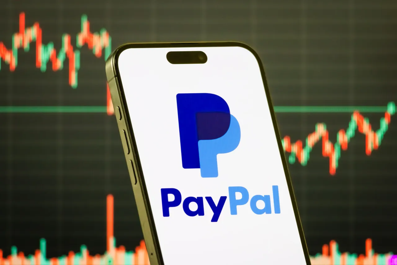 A smartphone displays the logo of PayPal Holdings in front of a screen showing a stock market chart. (Photo illustration by Cheng Xin/Getty Images)