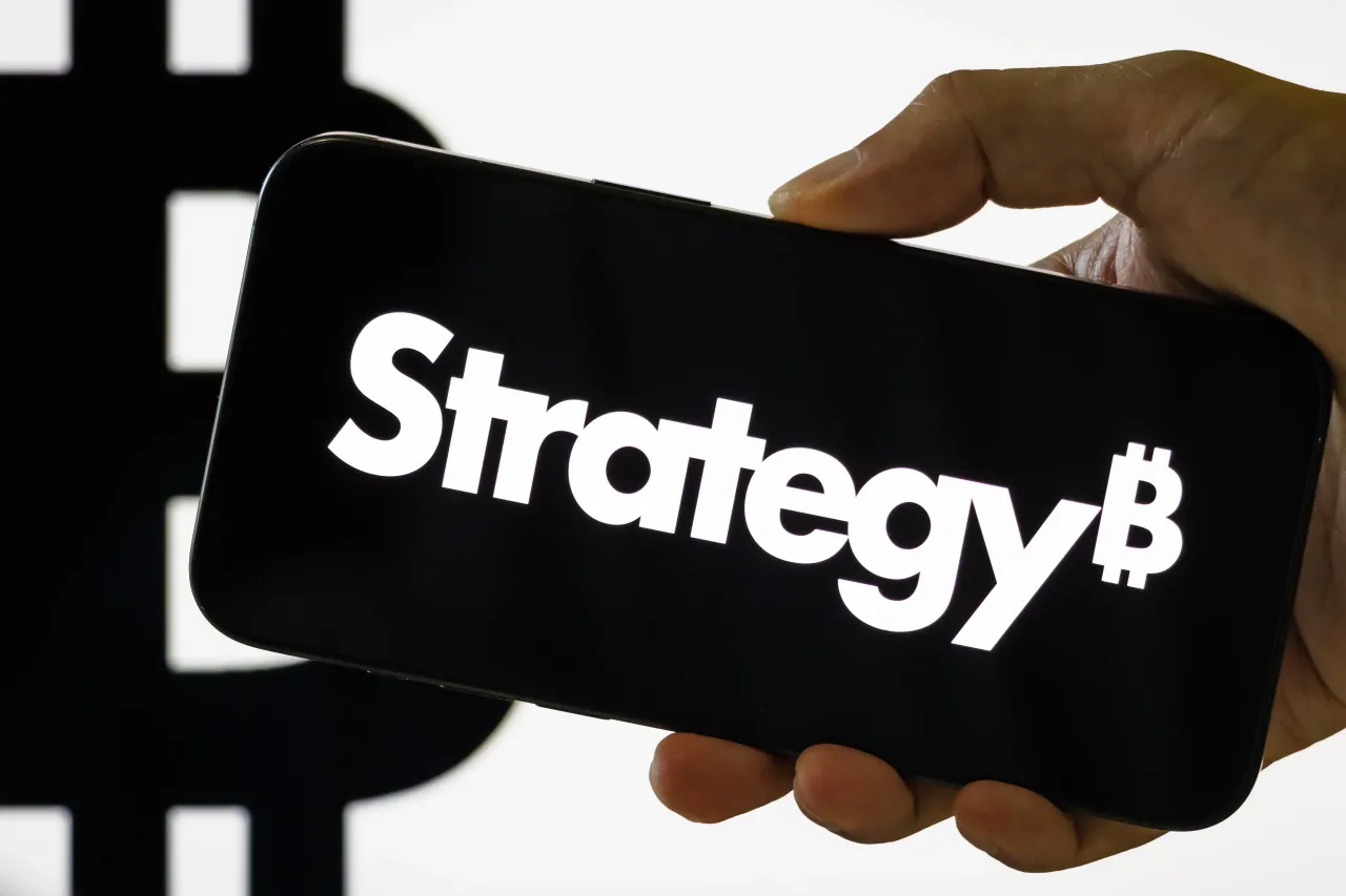 A person holds a smartphone displaying the stylized Strategy Inc. branding, with a large Bitcoin symbol visible in the background. Strategy stock and Bitcoin rallied last week. (Photo illustration by Cheng Xin/Getty Images)