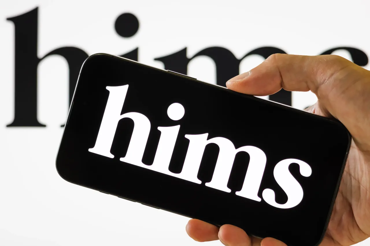 In this photo illustration, a person holds a smartphone displaying the logo of Hims & Hers Health Inc. (NYSE: HIMS). (Photo illustration by Cheng Xin/Getty Images)