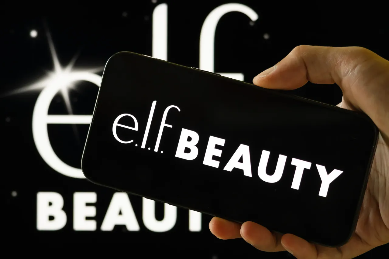 In this photo illustration, a person holds a smartphone displaying the logo of e.l.f. Beauty, in front of a screen showing the company's brand logo. (Photo illustration by Cheng Xin/Getty Images)