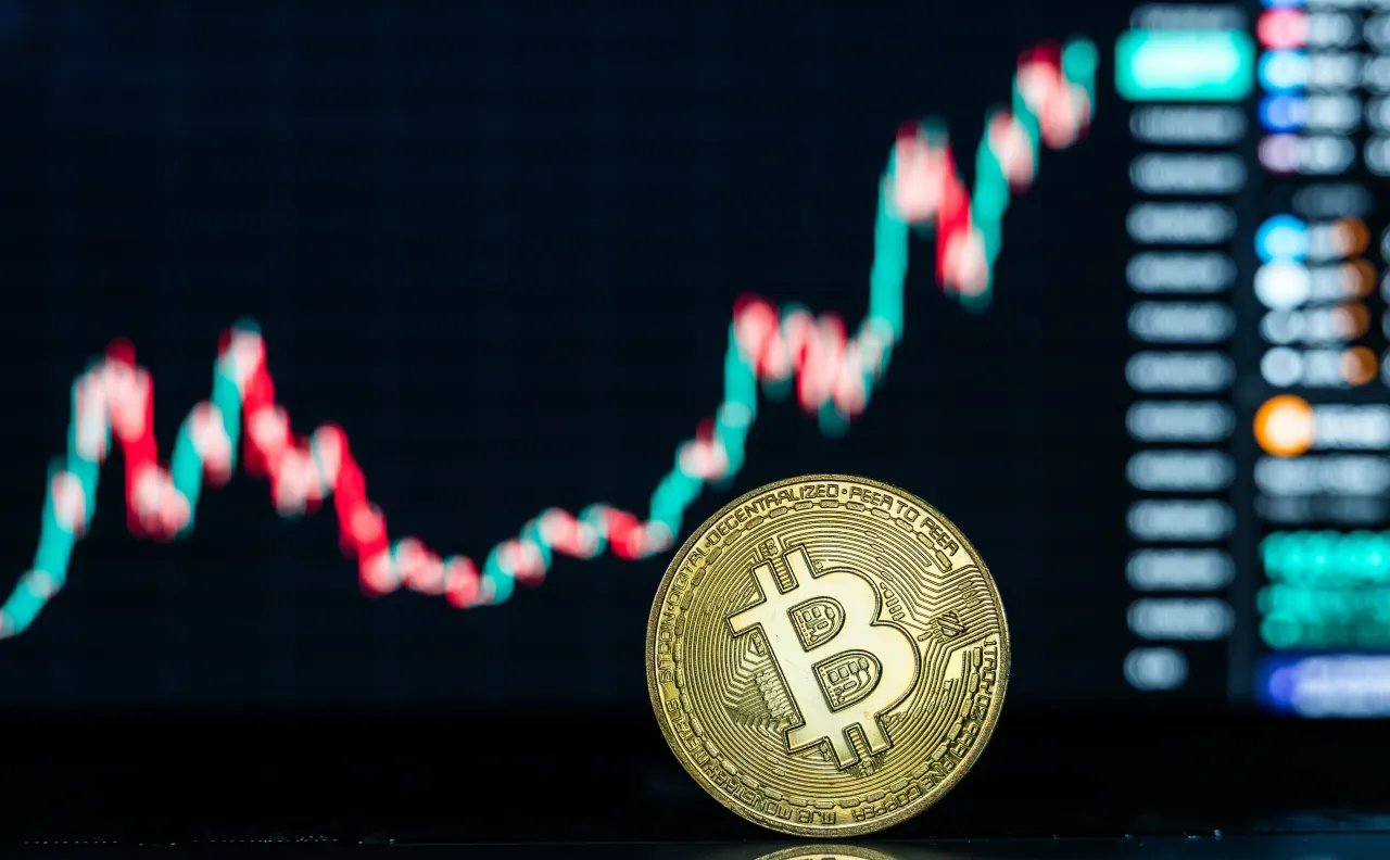 A symbolic Bitcoin coin can be seen on a laptop in front of a Bitcoin chart. 