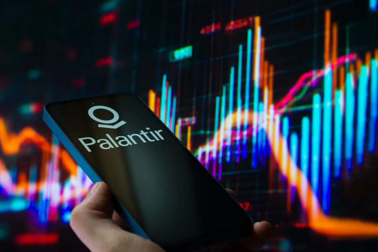 The Palantir logo is displayed on a mobile phone with a financial stock graph seen in the background. (Photo Illustration by Jonathan Raa/NurPhoto via Getty Images)