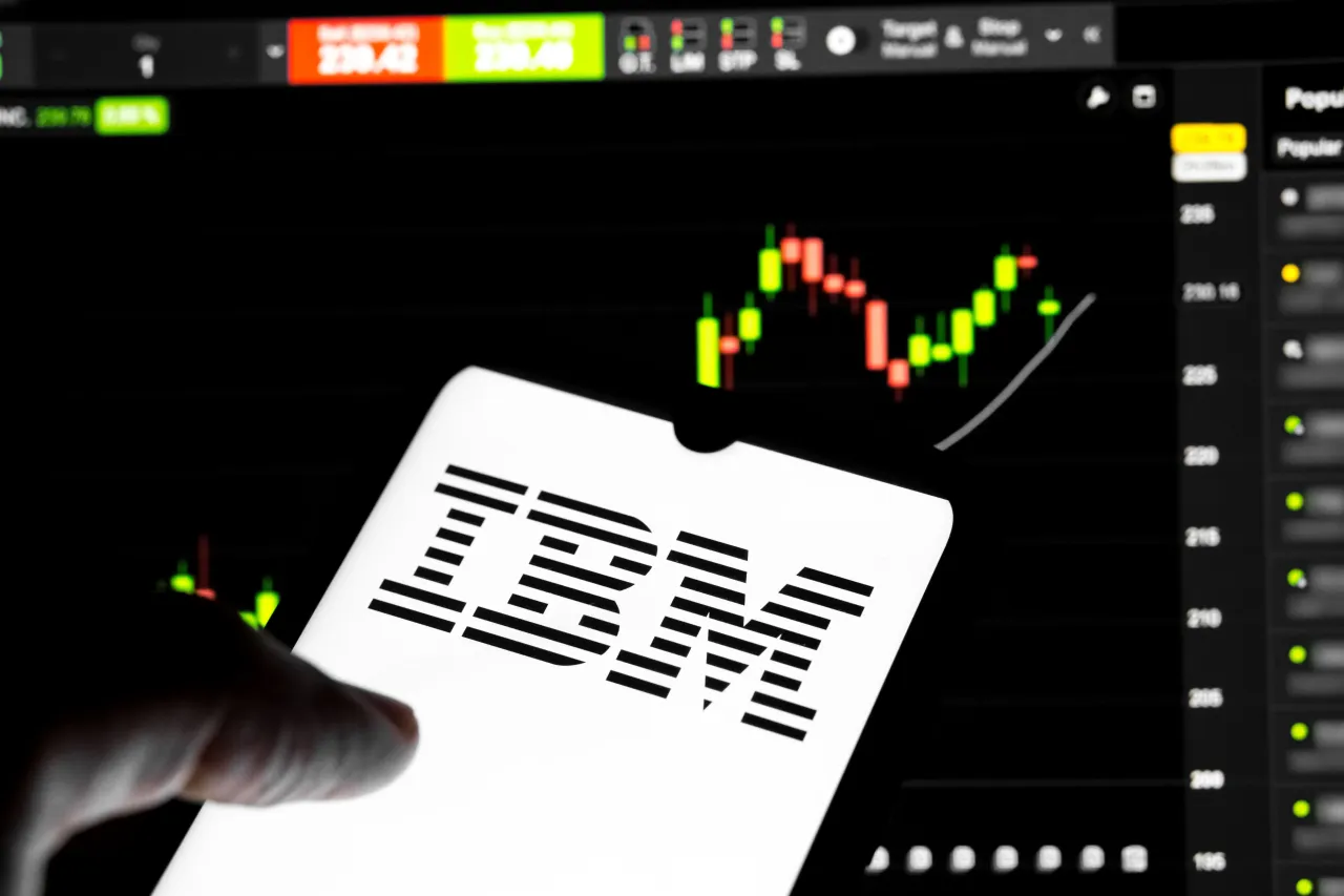 The IBM logo appears on a smartphone screen, and a stock exchange curve chart displays as the background. (Photo by Nikolas Kokovlis/NurPhoto via Getty Images)