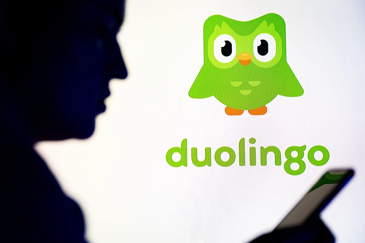 In this photo illustration, a silhouetted individual is seen holding a mobile phone with a Duolingo logo displayed in the background.