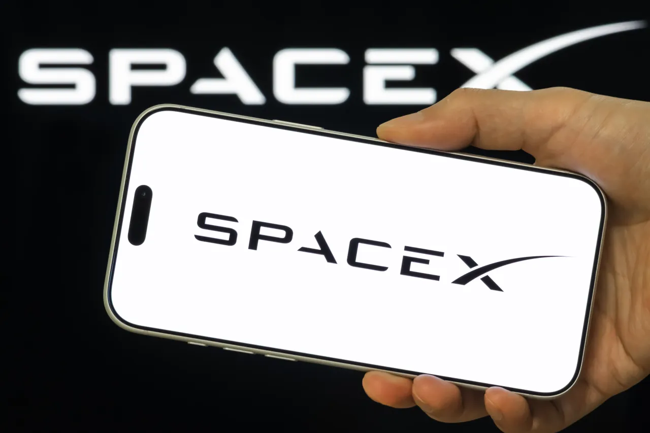 A person holds a smartphone displaying the SpaceX logo, with SpaceX branding visible in the background. (Photo illustration by Cheng Xin/Getty Images)