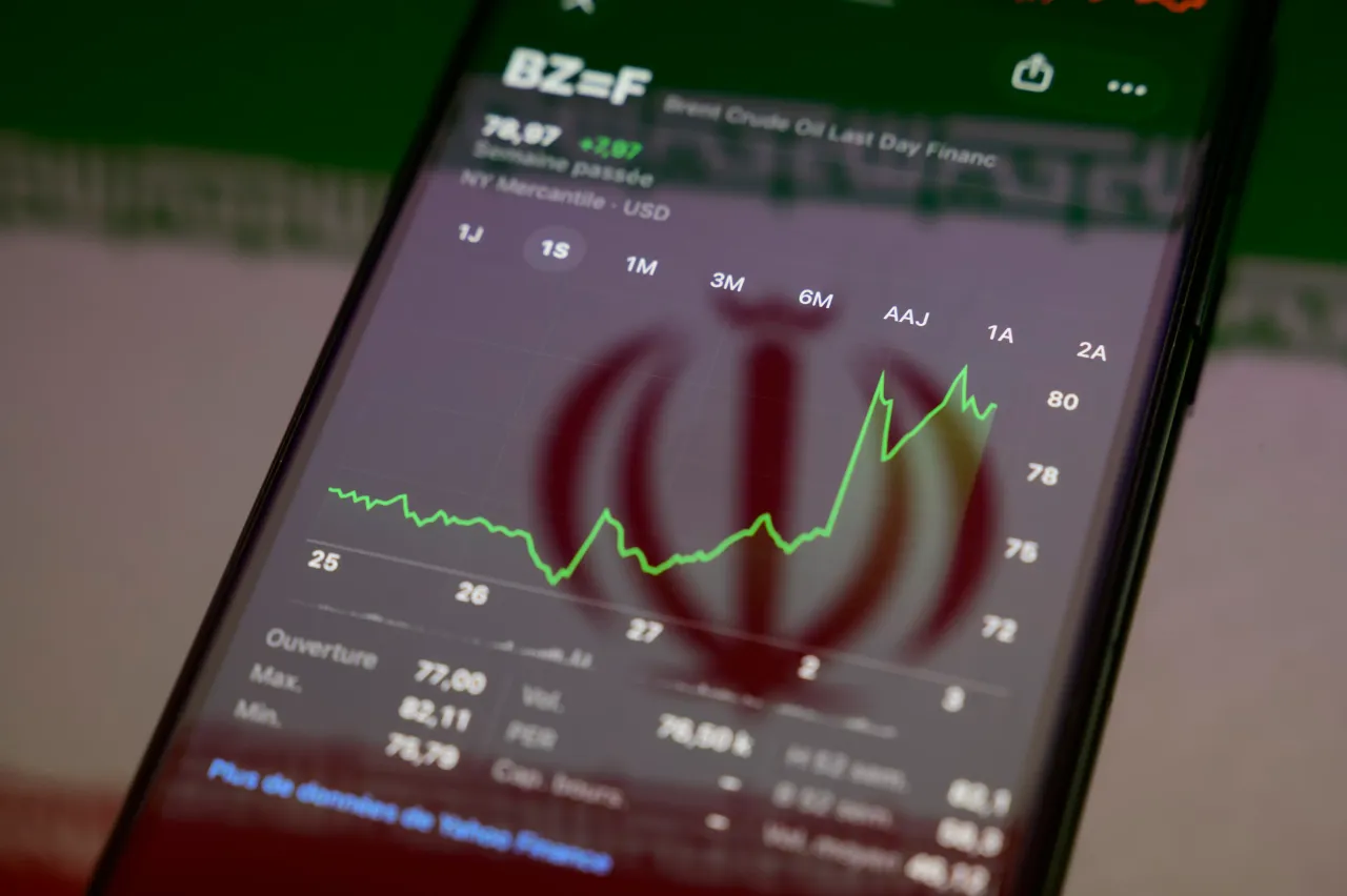 The Brent crude oil price chart is displayed on a smartphone screen placed on a reflective surface onto which an Iranian flag is projected. (Photo by Samuel Boivin/NurPhoto via Getty Images)