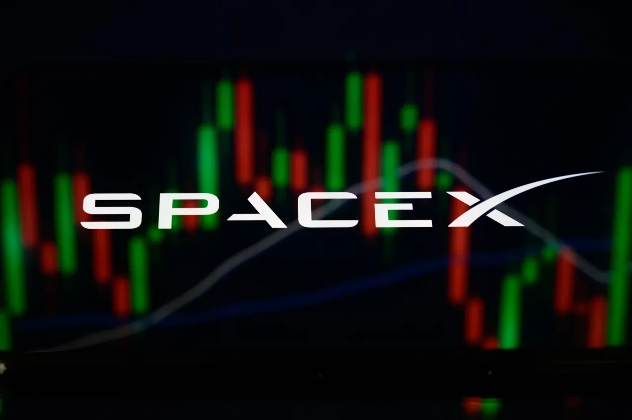 The SpaceX logo is displayed on the screen of a smartphone placed on a reflective surface onto which a stock market chart is projected. (Photo by Samuel Boivin/NurPhoto via Getty Images)