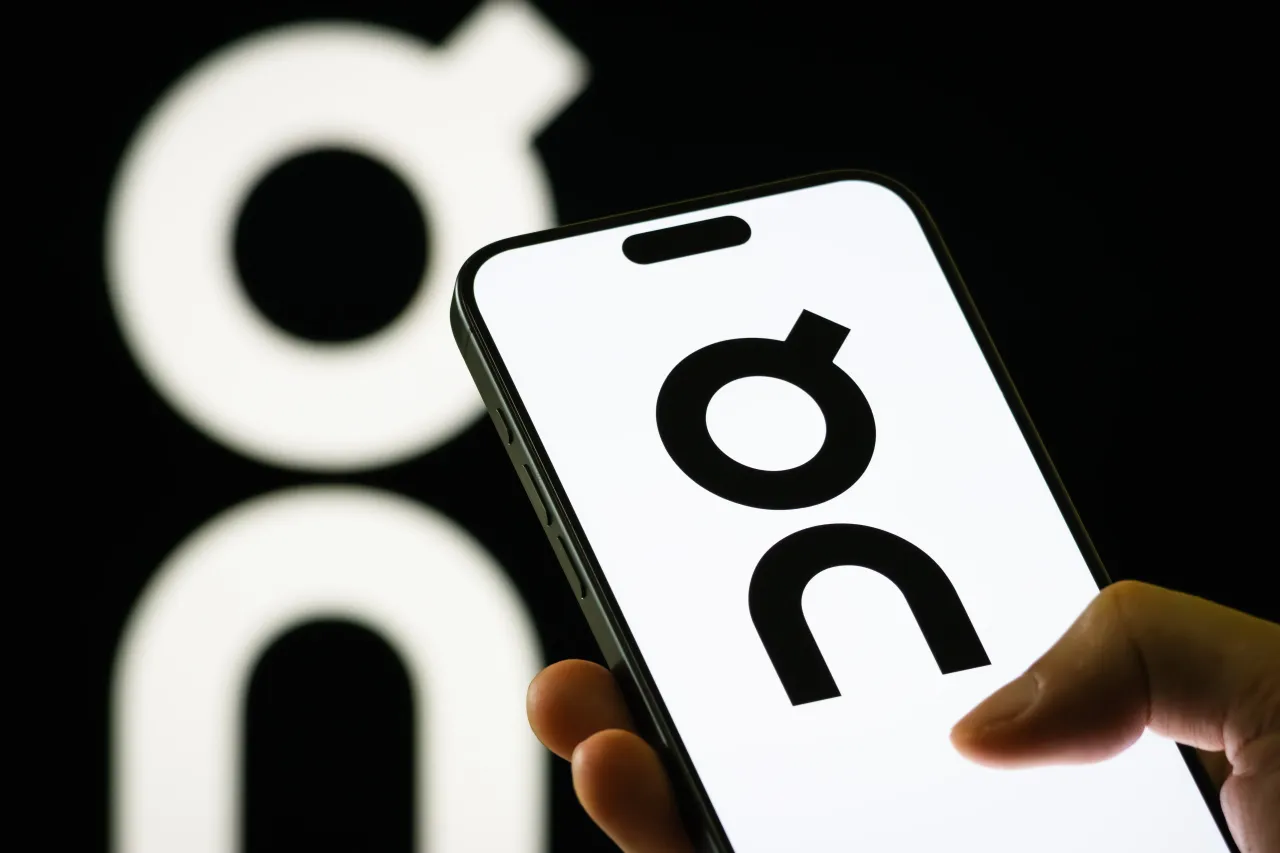 In this photo illustration, a person holds a smartphone displaying the logo of On Holding AG (NYSE: ONON), a Swiss sportswear company known for its high-performance running shoes and apparel. 