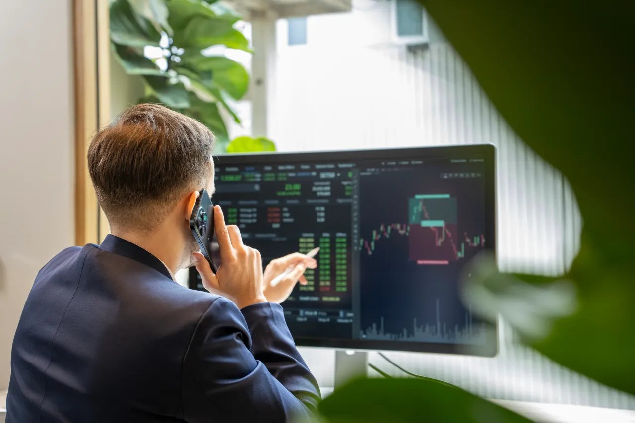 Stockbroker analyzing financial charts on a computer screen while engaging in a discussion about market trends with a client over the phone, demonstrating expertise in investment strategies. 