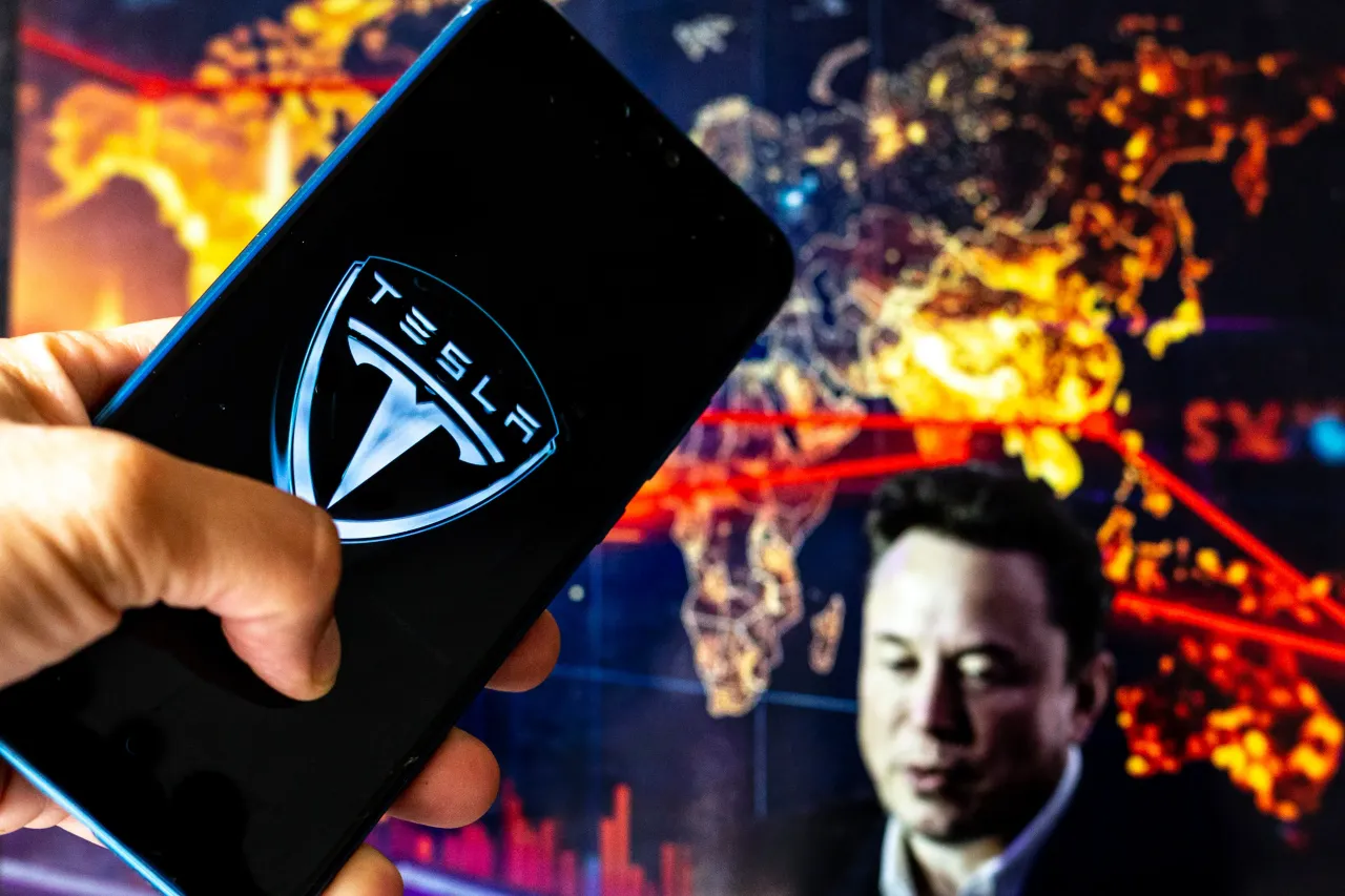 In this photo illustration, a Tesla logo is seen displayed on a smartphone with the image of Elon Musk in the background. 