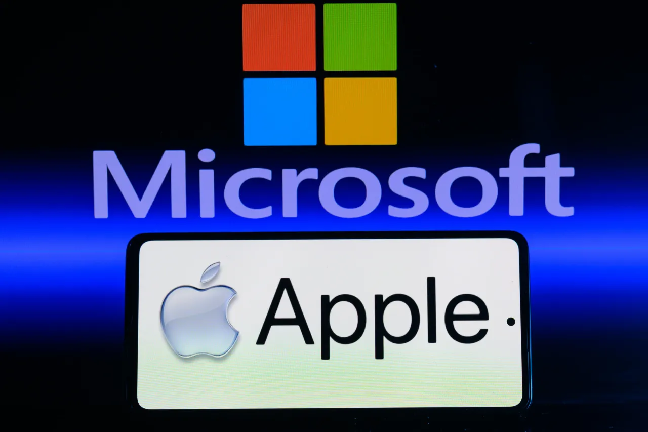 The Apple logo is being displayed on a smartphone with Microsoft visible in the background in this photo illustration