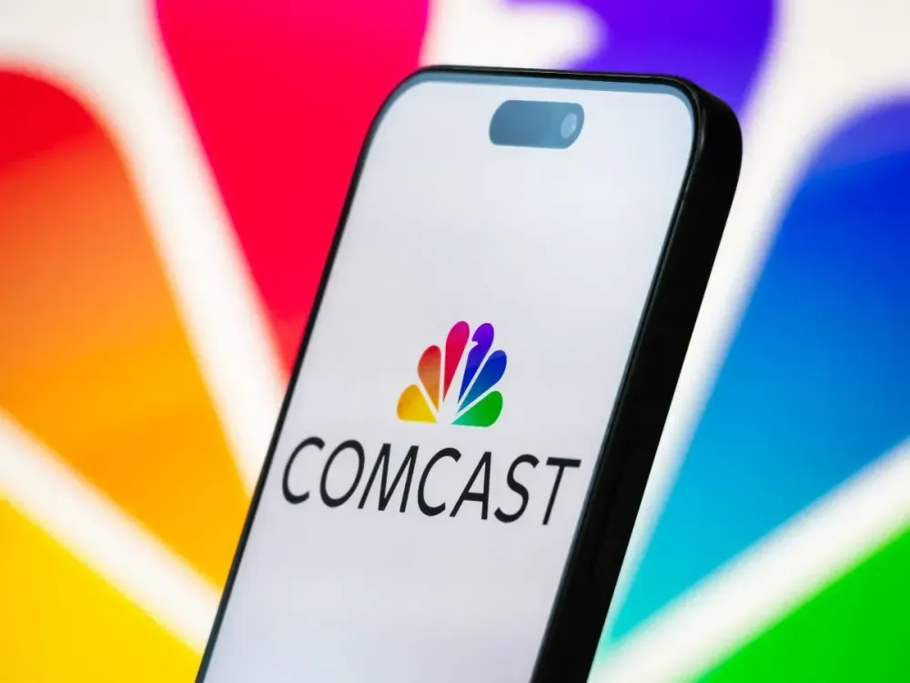 In this photo illustration, the Comcast logo is displayed on a smartphone screen, with the company's rainbow-colored NBC peacock branding visible in the background, on April 20, 2025, in Chongqing, China. (Photo by Cheng Xin/Getty Images)