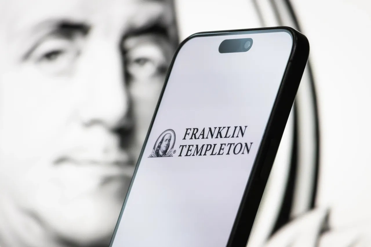 In this photo illustration, the logo of Franklin Templeton is displayed on a smartphone screen and Benjamin Franklin's portrait visible in the background, on April 26, 2025, in Chongqing, China. (Photo illustration by Cheng Xin/Getty Images)