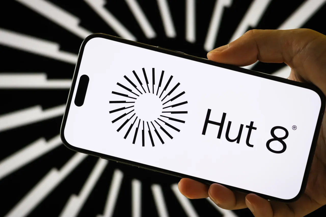 In this photo illustration, a person holds a smartphone displaying the logo of Hut 8 Corp. (HUT) in front of a screen showing the company's stylized black-and-white logo on August 7, 2025 in Chongqing, China. (Photo illustration by Cheng Xin/Getty Images)