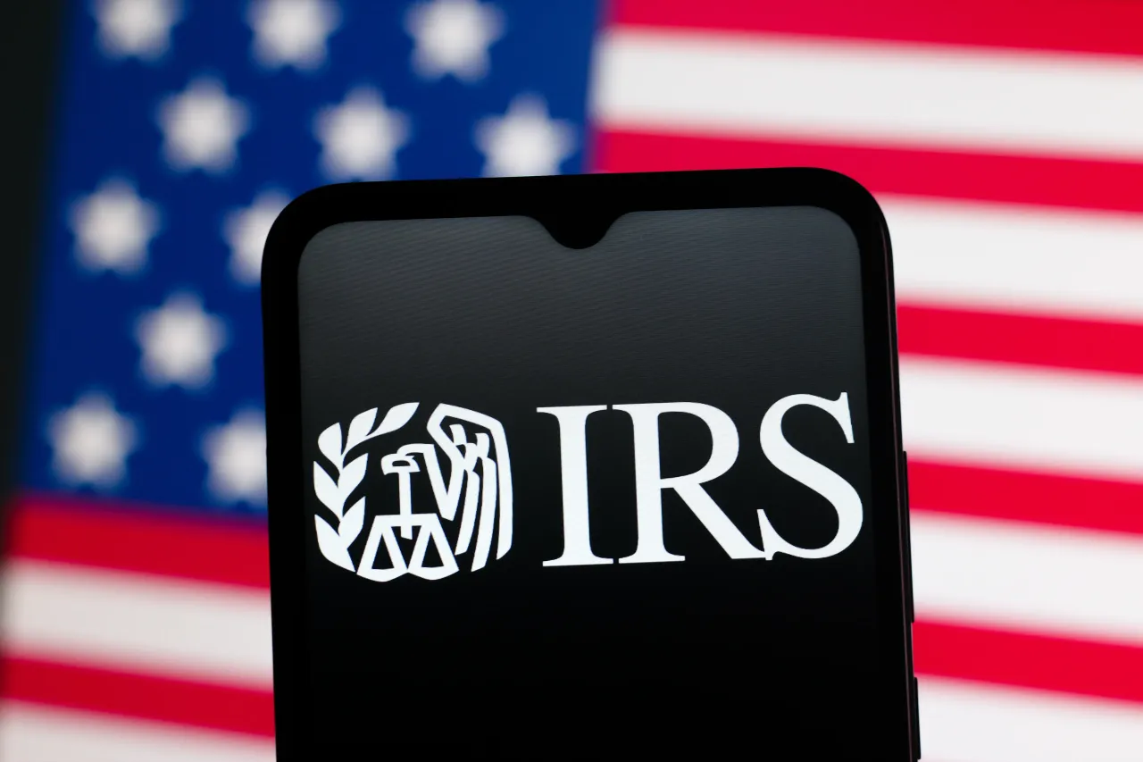 In this photo illustration, the Internal Revenue Service (IRS) logo is seen displayed on a smartphone screen and the United States flag in the background
