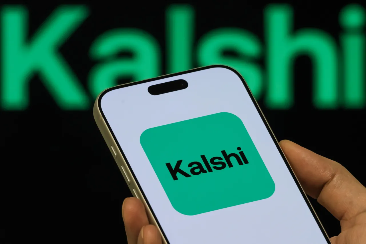 In this photo illustration, a person holds a smartphone displaying the Kalshi app icon, with the Kalshi branding visible in the background