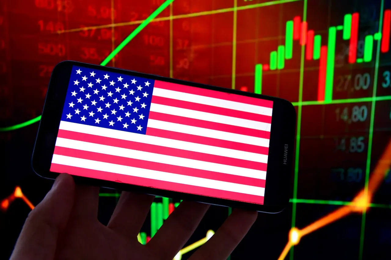 In this photo illustration, a US flag is displayed on a smartphone and stock market prices in the background. (Photo Illustration by Romain Doucelin/SOPA Images/LightRocket via Getty Images)
