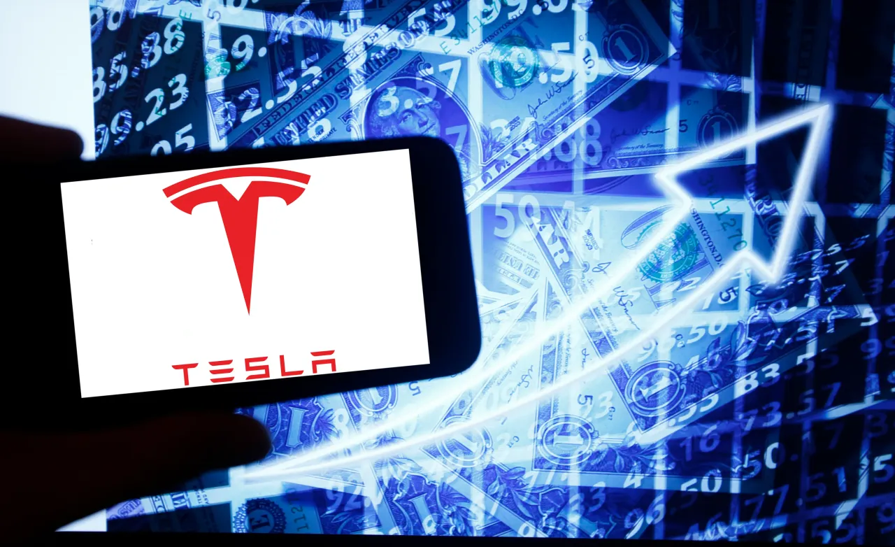 The logo of the Tesla Inc is seen on a screen of a smartphone next to a screen with an illustration ofthe stock market. (Photo by Alexander Pohl/NurPhoto via Getty Images)